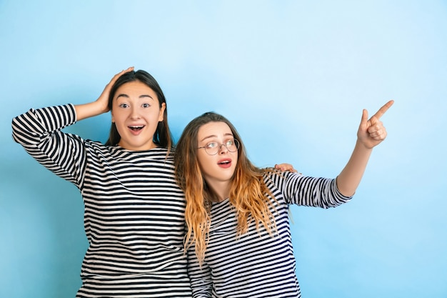 Zeigen erstaunt. Junge emotionale Frauen lokalisiert auf blauem Studiohintergrund der Steigung. Konzept der menschlichen Emotionen, Gesichtsausdruck, Freundschaft, Anzeige. Schöne kaukasische Modelle in Freizeitkleidung.