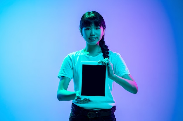 Zeigt den Bildschirm des Tablets an. Porträt der jungen asiatischen frau auf blau-violettem studiohintergrund der steigung im neonlicht. Konzept der Jugend, menschliche Emotionen, Gesichtsausdruck, Verkauf, Anzeige. Schönes brünettes Modell.