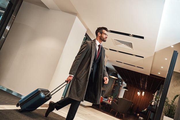 Zeit, dieses Hotel zu buchen. Menschen- und Technologiekonzept - glücklicher junger Mann mit Reisetasche, der vom Flughafen im Hotel spazieren geht. Untersicht