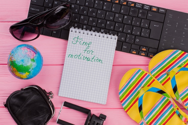 Zeit für die Motivation auf dem Notebook mit Brillenportemonnaie und mehrfarbigen Flip-Flops für Damen auf rosa Tischplattenhintergrund. Globus und schwarze Tastatur.