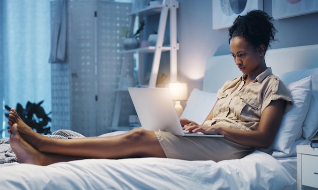 Zeit für ein nächtliches Bloggen Aufnahme einer attraktiven jungen Frau, die ihren Laptop benutzt, während sie nach einem langen Arbeitstag auf ihrem Bett liegt