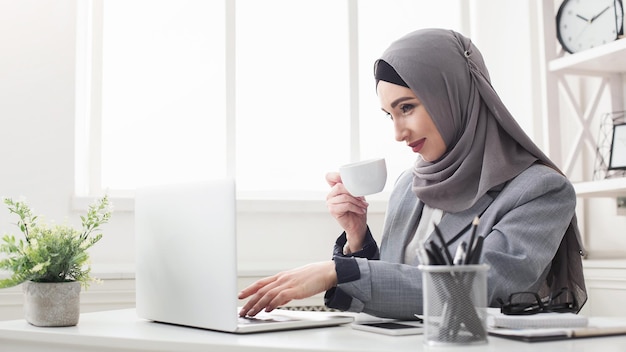 Zeit für Kaffee im Büro - Geschäftsfrau im Hijab, die arbeitet, Laptop benutzt und heißes Erfrischungsgetränk trinkt