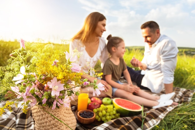 Zeit für Picknick. Glückliche liebevolle Familie, die auf Decke sitzt und spricht, selektiver Fokus auf Korb mit Blumen