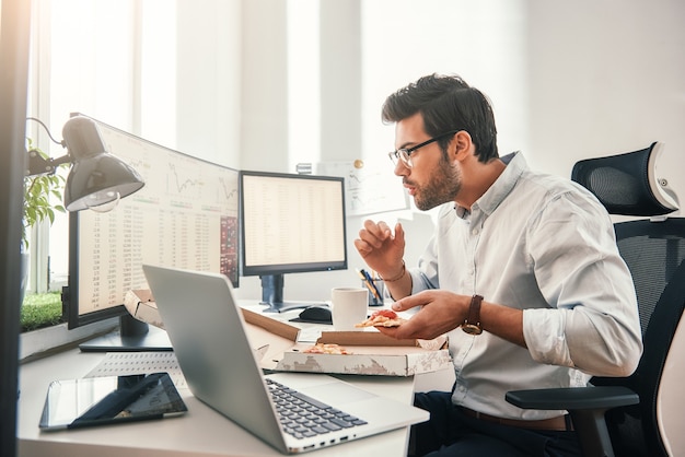 Zeit für Pizza Seitenansicht des jungen hungrigen Händlers isst heiße Pizza, während er in seinem modernen Büro auf den Bildschirm mit Finanzdaten schaut.