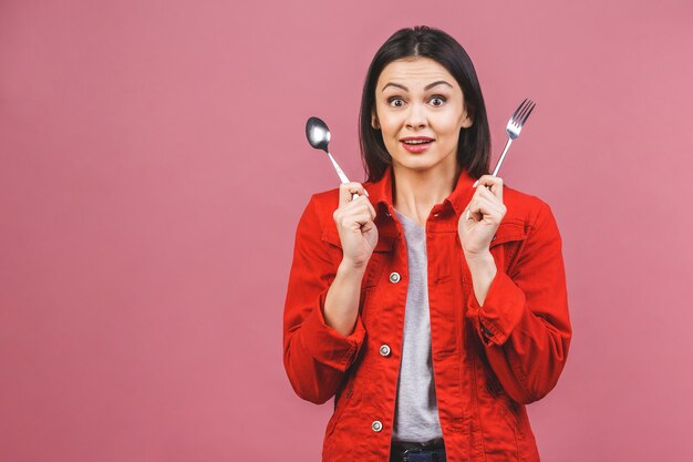 Zeit für's Abendessen! Porträt der jungen schönen Frau, die rotes Freizeithemd trägt, Löffel und Gabel lokalisiert über rosa Hintergrund hält.