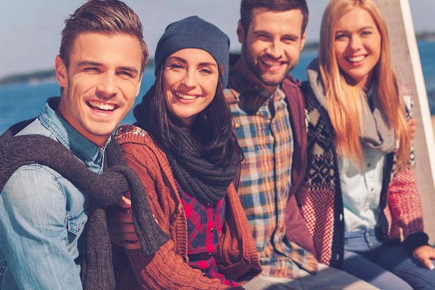 Zeit mit besten Freunden verbringen. Gruppe junger glücklicher Menschen, die sich miteinander verbinden und lächeln, während sie am Flussufer sitzen