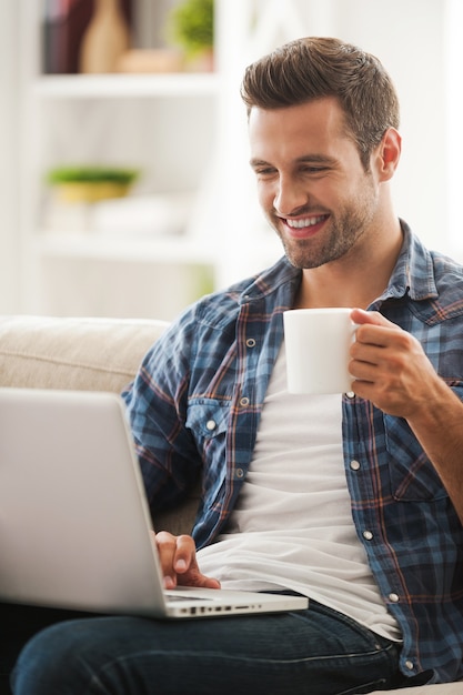 Zeit online verbringen. Lächelnder junger Mann, der am Laptop arbeitet und eine Tasse Kaffee hält, während er auf dem Sofa sitzt