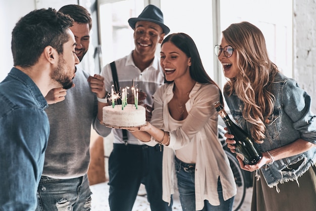 Zeit, sich etwas zu wünschen! Glücklicher junger Mann, der Kerzen bläst, während er Geburtstag unter Freunden feiert