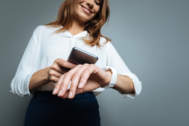 Zeit synchronisieren. Schöne entzückende hübsche Frau, die auf ihre Uhr schaut und lächelt, während sie Zeit mit ihrem Smartphone synchronisiert