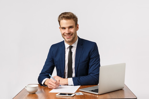 Zeit um Geld zu verdienen. Der schöne Bürovorsteher, der seinen Schreibtisch sitzt, Bericht und lächelnden antwortenden Kunden schreibt, Behälter, bereiten Dokumentation vor und verwenden Laptop, trinken Kaffee von der Schale