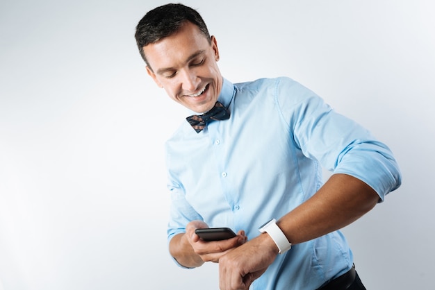 Zeit vergleichen. Positiv entzückter junger Mann, der auf seine Smartwatch schaut und sein Smartphone hält, während Zeit vergleicht