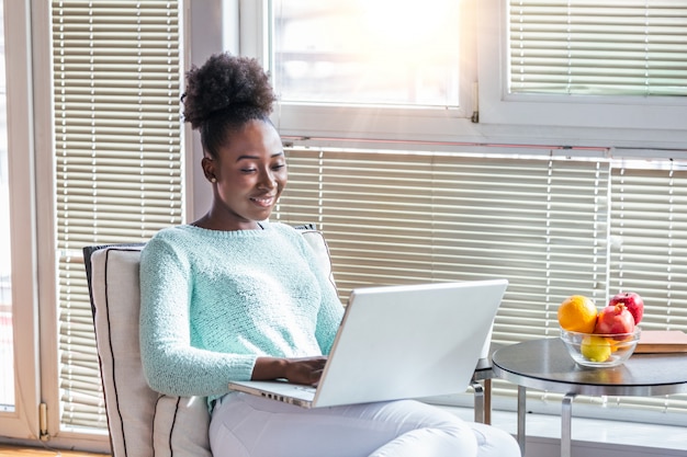 Zeit zu Hause genießen. Schöne junge lächelnde Frau, die an Laptop arbeitet und Kaffee beim in einem großen bequemen Stuhl zu Hause sitzen trinkt