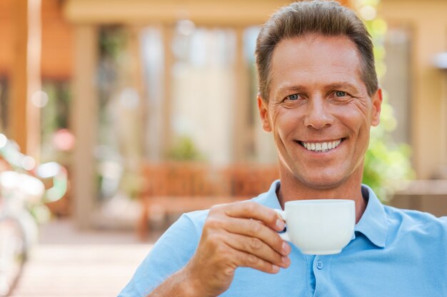 Zeit zum Entspannen. Fröhlicher reifer Mann, der Kaffee trinkt und lächelt, während er draußen mit Haus im Hintergrund sitzt