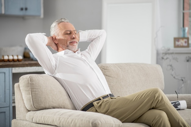 Zeit zum Träumen. Positiv entzückter bärtiger Mann, der Beine überkreuzt, während er auf dem Sofa sitzt und Arme streckt