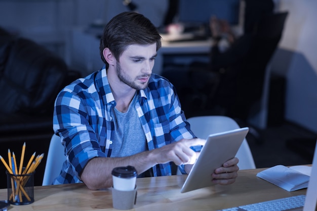 Zeitalter der Technologie. Hübscher junger bärtiger Mann, der am Tisch sitzt und Tablette beim Kaffee verwendet