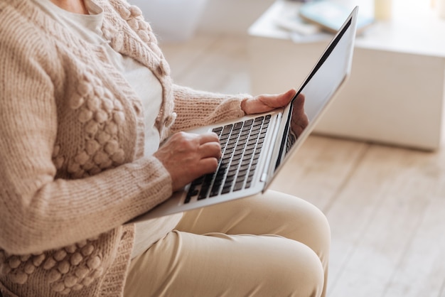 Zeitalter der Technologie. Schließen Sie oben von einem modernen innovativen Laptop, der in den Händen einer angenehm netten älteren Frau ist, während Sie daran arbeiten