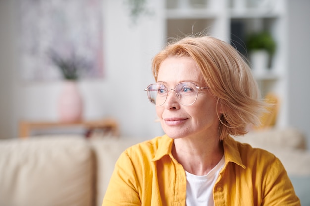 Zeitgenössische blonde reife Frau in gelber Freizeitkleidung und Brille, die Gelassenheit ausdrückt, während sie sich in der häuslichen Umgebung ausruht