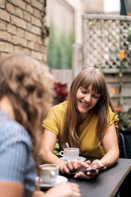 Zeitgenössische Freundinnen genießen Kaffee am Tisch im Café, während sie Smartphone teilen und lächeln