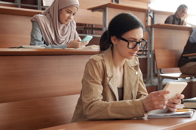 Zeitgenössische junge interkulturelle Studierende, die im Hörsaal an Schreibtischen sitzen und in mobilen Geräten scrollen, während sie nach Online-Daten suchen