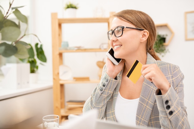 Zeitgenössische mobile Frau in der Abendgarderobe, die mit Manager des Online-Shops auf Smartphone spricht, während Bestellung im Büro in der Pause macht