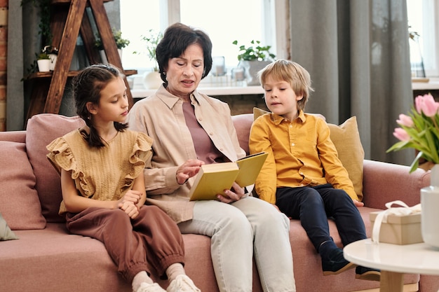 Zeitgenössische reife brünette Frau in Freizeitkleidung, die auf der Couch zwischen ihren süßen kleinen Enkelkindern sitzt und ihnen Märchen vorliest
