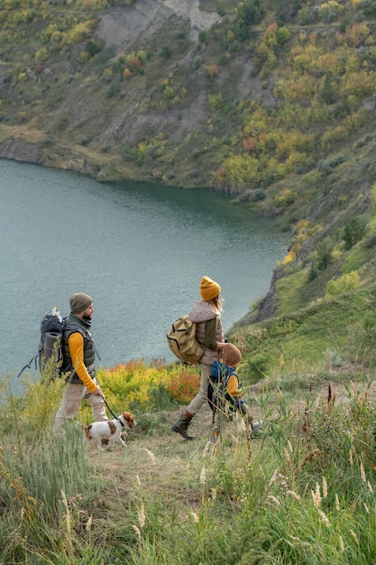 Zeitgenössische Rucksacktouristen in warmer Freizeitkleidung, die sich auf einem Fußweg entlang eines Flusses oder Sees bewegen, während sie einen Wochenendausflug in der natürlichen Umgebung genießen