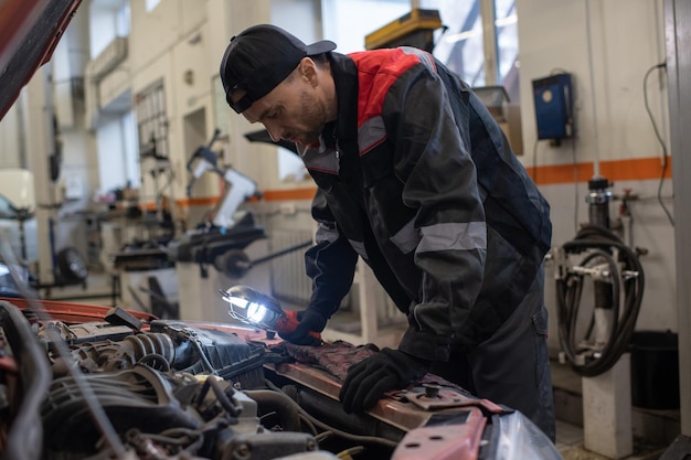 Zeitgenössischer Automechaniker in Arbeitskleidung, der sich über die offene Motorhaube beugt