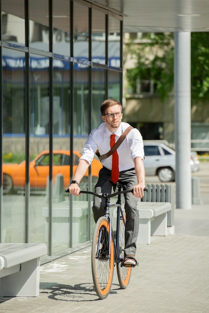 Zeitgenössischer junger Geschäftsmann, der Fahrrad an sonnigem Sommertag reitet und sich außerhalb des Geschäftszentrums auf seinem Weg zur Arbeit bewegt