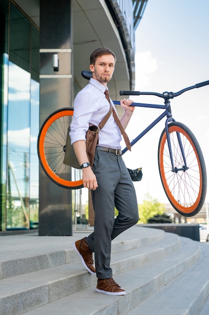 Zeitgenössischer junger Geschäftsmann, der sein Fahrrad trägt, während Bürogebäude verlässt und unten gegen Äußeres des Bürozentrums bewegt