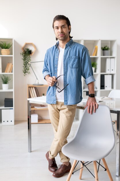 Zeitgenössischer junger Geschäftsmann in der Freizeitkleidung, die durch Stuhl vor Kamera mit Schreibtisch auf Hintergrund steht