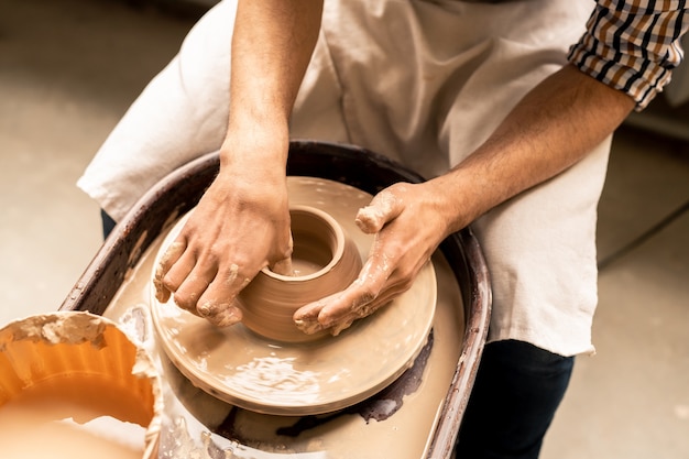 Zeitgenössischer junger Meister der Keramik, der durch rotierendes Rad sitzt, während Topf aus flüssigem Ton in Werkstatt formt