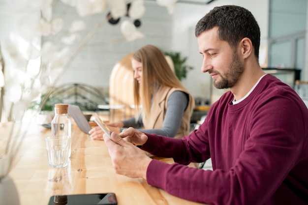 Zeitgenössischer junger mobiler Mann, der in digitaler Tablette rollt, während Ruhe durch Tisch im Café hat