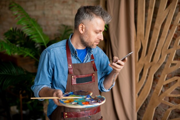 Zeitgenössischer kreativer Maler mit Palette und Pinsel, die in Smartphone oder Nachrichten im Studio scrollen