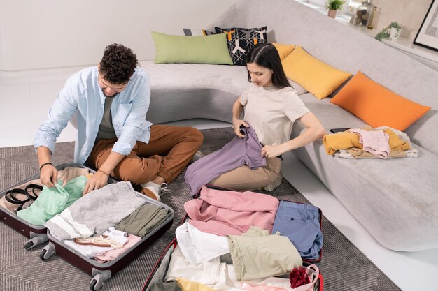 Zeitgenössisches junges Paar in Freizeitkleidung, das auf dem Boden neben einer großen Couch sitzt und seine Kleidung packt, während es auf die Reise geht