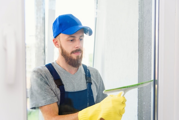 Zelador masculino usando um rodo de borracha para limpar uma janela em um escritório usando um avental e luvas enquanto ele trabalha