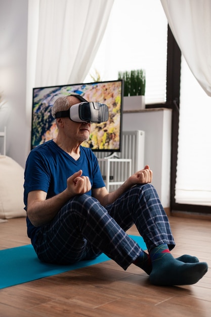 Zen Senior Mann sitzt auf Yogamatte im Lotussitz während des Pilates-Trainings im Wohnzimmer und streckt den Körpermuskel. Rentner mit Virtual-Reality-Headset übt Meditation aus