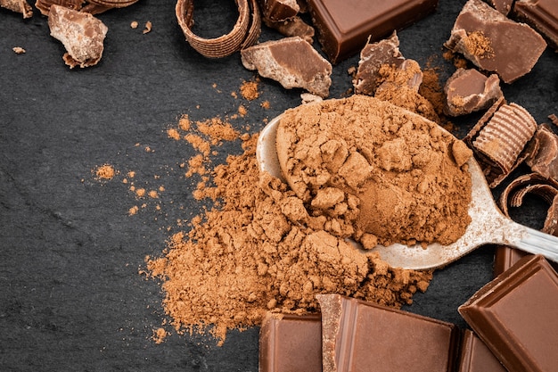 Zerbrochene Schokoladenstücke und Kakaopulver auf einer dunklen Oberfläche.