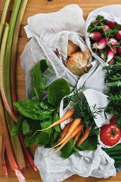 Zero Waste Lebensmitteleinkaufskonzept Flache Lage Wiederverwendbare umweltfreundliche Taschen mit frischem Gemüse, Karotten, Tomaten, Spinacharugula, Pilzen, Rhubarbonions auf Holztisch, verbieten Kunststoff