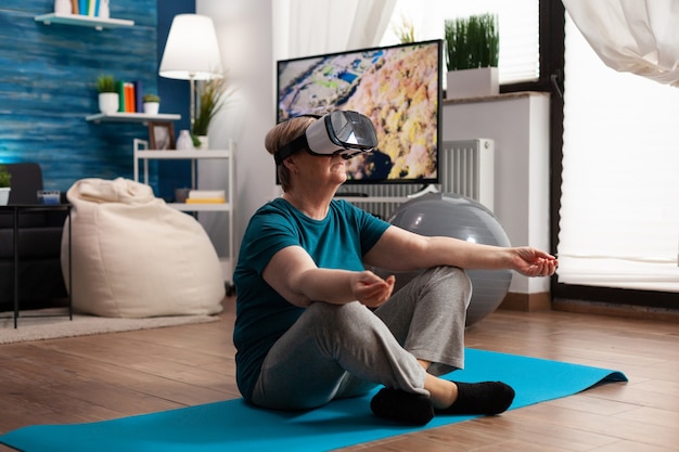 Ziehen Sie eine ältere Frau zurück, die während des Pilates-Meditationstrainings auf einer Yogamatte im Lotussitz im Wohnzimmer sitzt und ein Virtual-Reality-Headset trägt