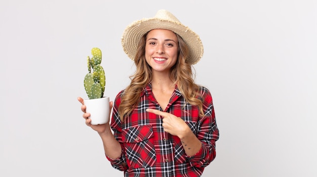 Ziemlich dünne Frau, die fröhlich lächelt, sich glücklich fühlt und auf die Seite zeigt, die einen Kaktus hält. Landwirt-Konzept