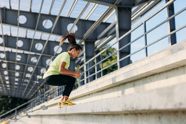 Ziemlich fitte Frau in der Sportkleidung, die auf Treppen am modernen Stadion springt.