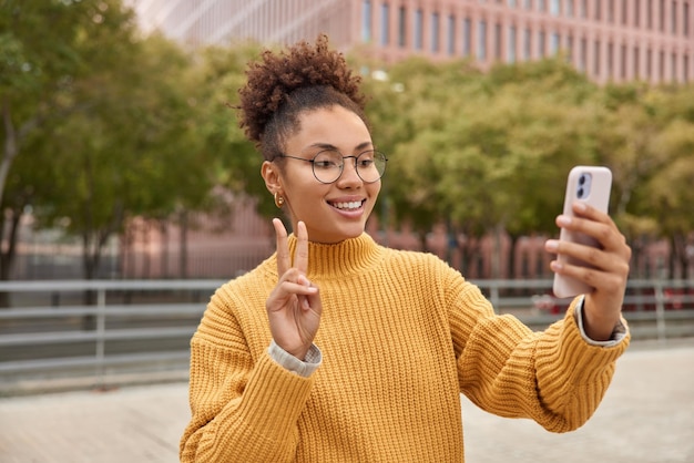 Ziemlich fröhliches tausendjähriges Mädchen trägt eine runde Brille und ein gelber Strickpullover macht Friedenszeichen, um Selfies vor der Kamera von Smartphone-Ständern im Freien in der Stadt zu machen Moderner Lebensstil