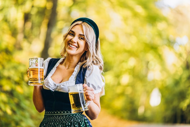 Ziemlich glückliche Blondine im Dirndl, traditionelles Festkleid, zwei Krug Bier draußen im Wald haltend