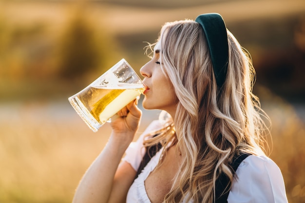 Ziemlich glückliche Blondine in Dirndl, traditionelles Festkleid, Bier draußen auf dem Feld trinkend