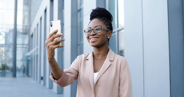 Ziemlich glückliche junge afroamerikanische junge Frau in den Gläsern, die Videochat auf Smartphone im Freien am Geschäftsgebäude haben. Freudige schöne Frau, die über Webcam auf Handy spricht und Videochatting.