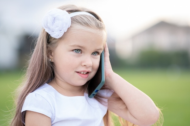 Foto ziemlich glückliches kindermädchen, das im sommer am handy lächelt und draußen spricht.