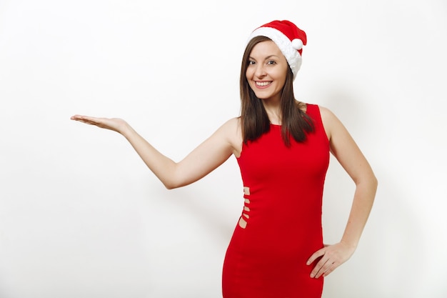 Ziemlich kaukasische glückliche Frau mit charmantem Lächeln mit rotem Kleid und Weihnachtsmütze, die Kamera hält und Kopienraum auf der Handfläche hält, isoliert auf weißem Hintergrund. Sankt-Mädchen. Neujahrsfeiertag 2018 Konzept.