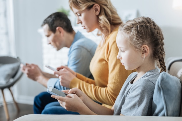 Ziemlich konzentriertes blondes kleines Mädchen, das ihr Telefon hält und ansieht, während es ordentlich bei ihren Eltern sitzt und sie auch ihre Telefone benutzen