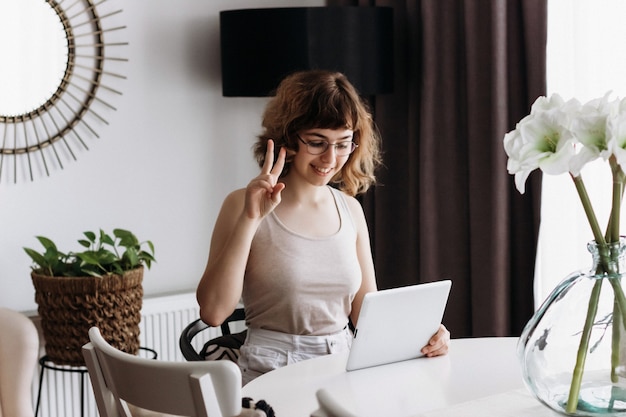 Ziemlich lockiges lächelndes junges Mädchen, das zu Hause am Tisch telefoniert. Videochat mit Freunden