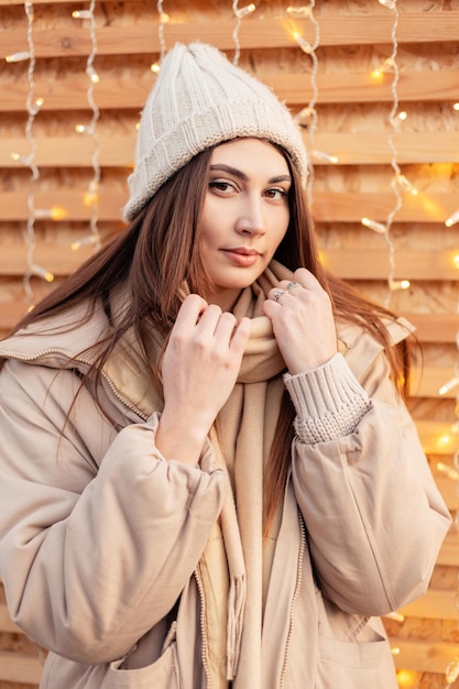 Ziemlich modisches junges Mädchen in stylischer Winteroberbekleidung mit Strickmütze und Jacke steht in der Nähe einer Holzwand mit Lichtern